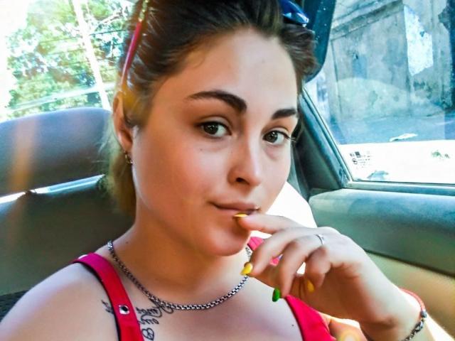 NanalyBeautyy, con la mano vicino alla bocca, indossa una maglietta rossa, ha le unghie verdi e gialle, un anello, una collana e un piccolo tatuaggio sul petto, seduta in un'auto..