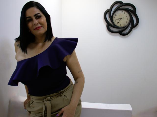 RebekaHunter has straight black hair, wearing a deep purple off-shoulder top with a ruffled hem, and beige pants with a tied belt. She has a tattoo on her right shoulder and another small tattoo on her left arm. She stands slightly leaning forward, smiling with red lipstick. A black, flower-shaped clock with white face and black numbers is on the white wall behind her. The lighting is even, highlighting her dark hair and purple top. Her expression is confident.