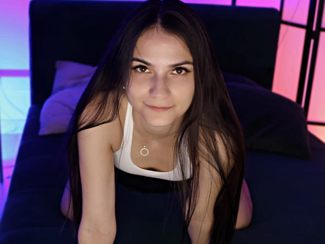 BabeRia with long black hair, wearing a white tank top, kneeling on a black bed, looking up with a subtle smile, small tits, cute face, pink and blue lighting.