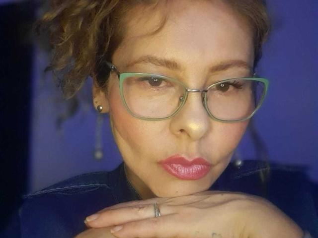 MiaAmoretti with curly brown hair, green glasses, pink lipstick, and a pensive expression, holding her chin with a ring on her hand. Her ass is not visible.