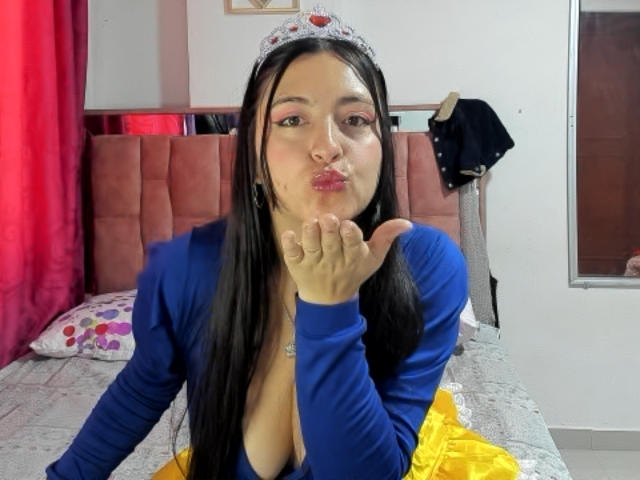 VenusSweetPalmer with long black hair, blue top, and tiara, pouting with her finger to her lips, showing her tits in a yellow skirt, on a bed.