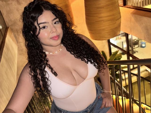 AprielKeith with curly black hair, pale pink tank top showing big tits, blue denim shorts, and a seashell necklace, smiling in a warmly lit, indoor setting.