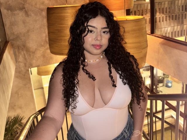 Apriel Keith tiene el cabello negro y rizado, escote rosa, y lleva una camiseta rosa con jeans de cintura alta, además de un collar de cuentas, y está de pie en una habitación con una iluminación cálida..