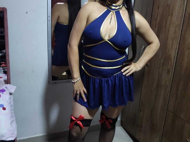 VenusSweetPalmer in a blue and gold halter dress with a cleavage cutout, black fishnet stockings with red bows, standing with hands on hips, long black hair.