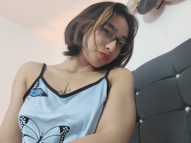 CamiVargas, con cabello corto, negro y rubio, gafas, piercing en la nariz y lápiz labial rojo, vestida con un camisón azul claro con diseño de mariposa, mostrando pequeños senos y un tatuaje de mariposa..