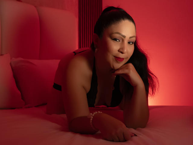 ArabelaFoxy is lying on a bed, wearing black lace lingerie, with red lighting, her hand resting on her chin, and a subtle smile on her face. Her tits are visible.