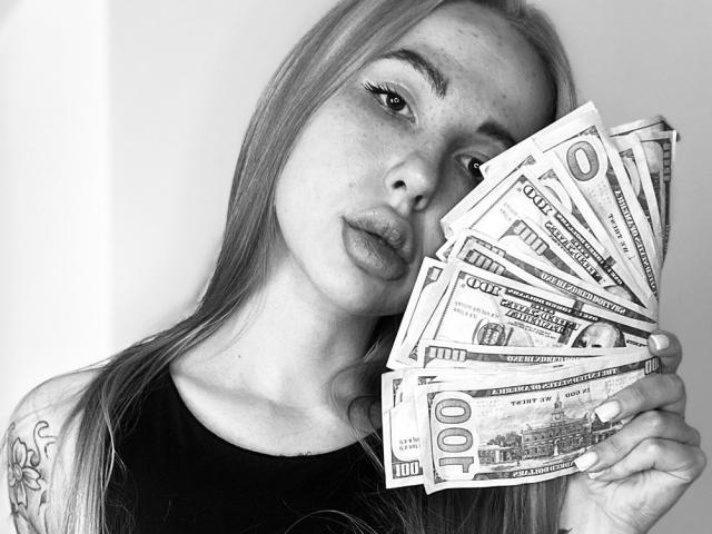 AmiraaGold with long blonde hair, holding a fan of dollar bills, pouty lips, tattoo on shoulder, black shirt, white background, black and white photo.