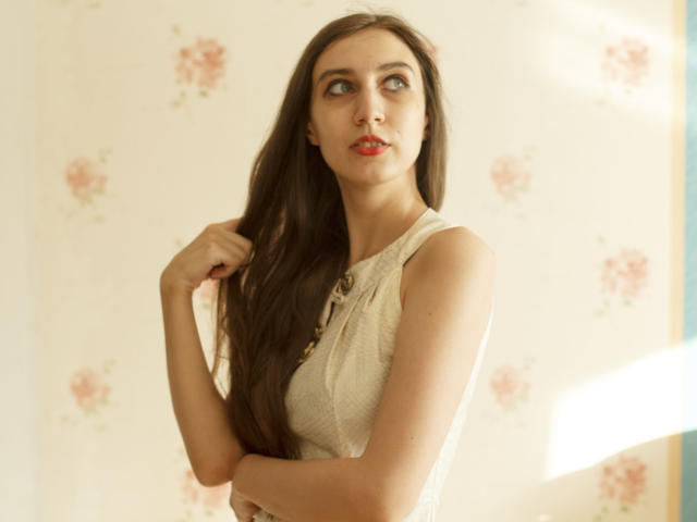 SummerCathy has long brown hair, red lipstick, and a white sleeveless top with buttons. She's looking up with a slight smile, touching her hair with her right hand.
