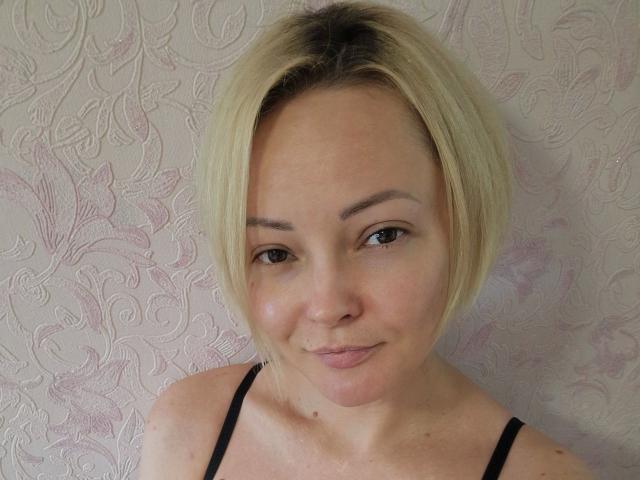 SophieCrush-hot with blonde bob, light skin, small nose, thin eyebrows, black tank top, subtle smile, cute face, small tits, standing against floral-patterned wall, head and shoulders shot.