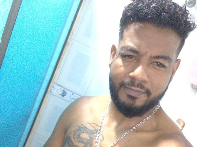 BladeFire, shirtless with a dark beard and short curly hair, wearing a chain necklace, showing a chest tattoo, smiling at the camera. Blue wall on the left.
