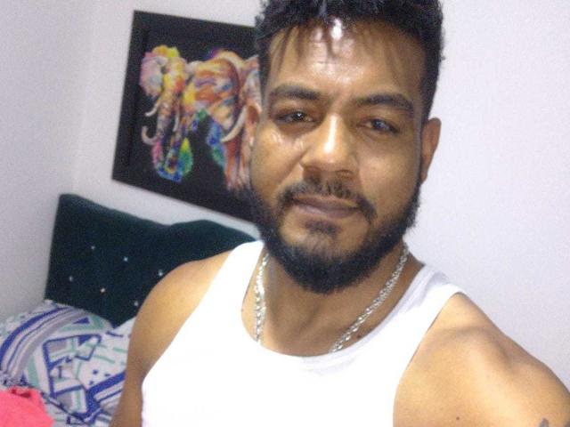 BladeFire, a muscular dude with dark skin and a short beard, wearing a white tank top and silver chain, smiling at the camera, with a colorful elephant painting behind.
