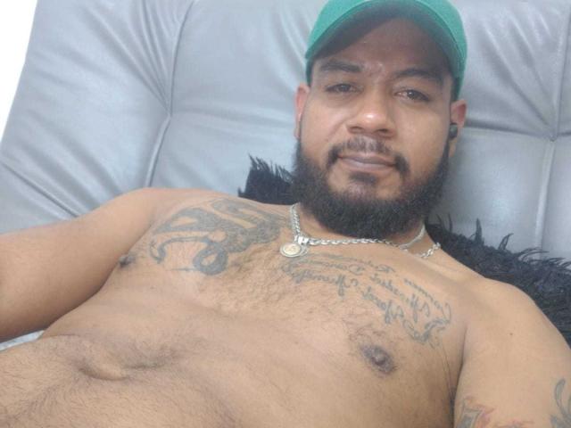 BladeFire, a muscular, bearded man with tattoos, green cap, and chain necklace, lying shirtless on a gray leather couch, showing his hairy chest and belly.