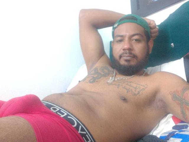BladeFire, shirtless with tattoos, wearing red boxer briefs showing a bulge, green cap backward, arm raised, looking at camera, chain necklace, slight beard, relaxed on bed.