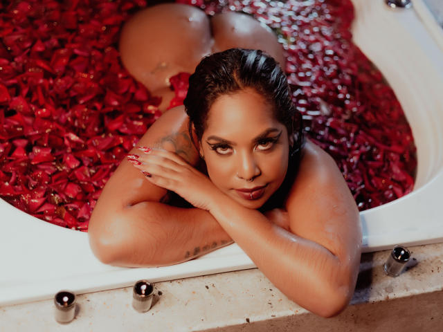 MadisonHetfield, laying in a bathtub filled with red rose petals, looking up with wet, dark hair, red nails, and a tattoo on her right arm, her ass above water.