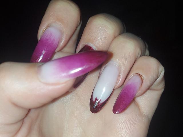 Close-up of JuliaHuntis's hand showing long, square-tipped nails with a gradient from dark purple at the tips to white at the bases. The nails have a glossy, shiny finish. The fingers are slightly curled, highlighting the nails' length and shape. The skin tone is light, and the nails are well-manicured. The background is dark, making the nails' colors stand out. The image focuses on the nails, with no other elements visible.