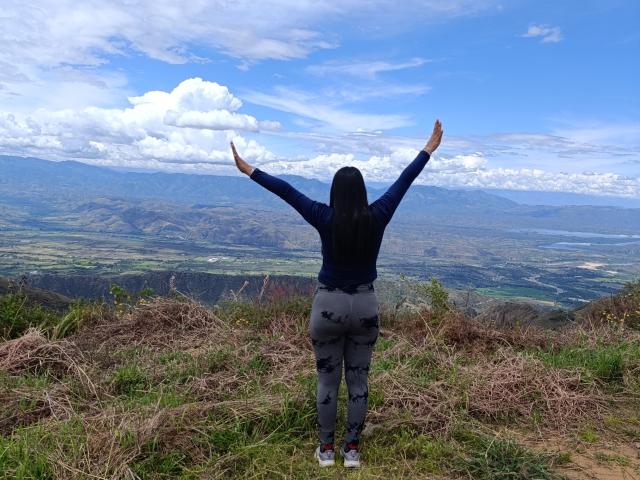 PauletteLoveX stands with arms raised, facing mountains, wearing a blue long-sleeve top and gray patterned leggings, on a grassy, rocky hill under a bright blue sky.