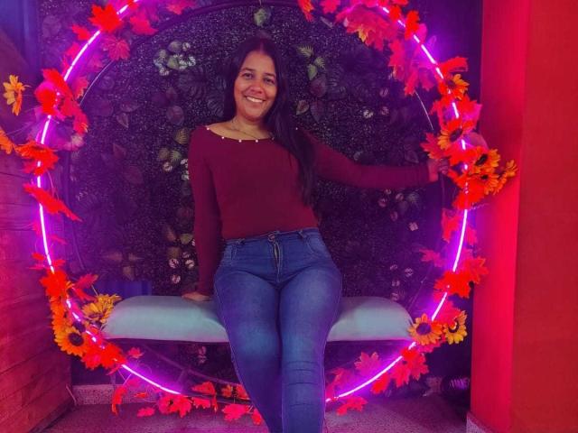 PauletteLoveX, sitting on a swing with purple neon lights and red-orange flowers, wearing a red top, blue jeans, smiling, showing her cute ass, long dark hair.