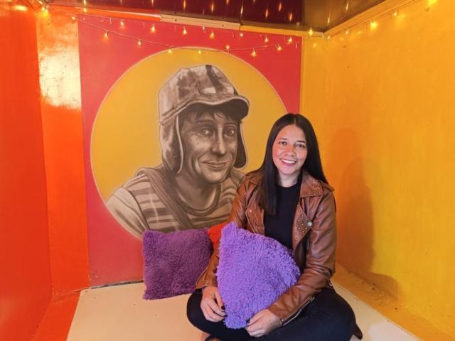 PauletteLoveX with long black hair, smiling, wearing a brown leather jacket, black top, and black pants, sitting on the floor, holding purple and purple fluffy pillows.