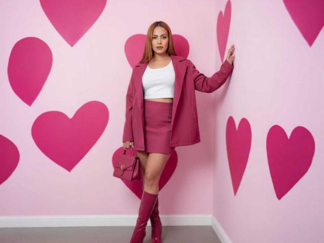 SharomCastle with pink hearts, wearing a pink jacket, white crop top, pink skirt, and matching pink boots, holding a pink handbag, standing against a pink wall.