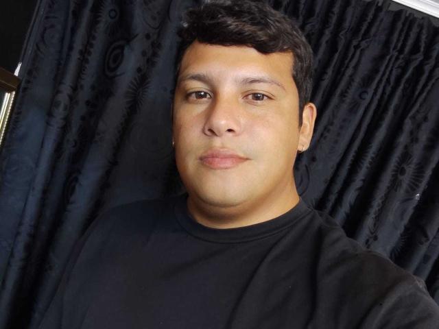 JustinBerry has a chubby face, short black curly hair, brown eyes, and is wearing a black shirt. His skin is light brown and he's giving a slight smile.