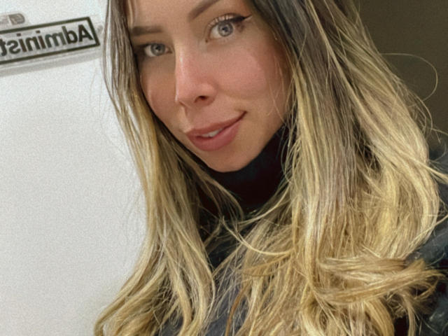 JennyLawry has blonde, wavy hair, light skin, and a cute smile. She's wearing a black turtleneck, and her blue eyes are slightly open. She's looking at the camera.