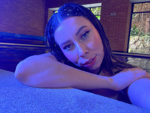 JennyLawry with wet black hair, blue-tinged skin, and eyeliner, resting her arm on a pool edge, looking at the camera with a slight smile, showing her bare shoulders.