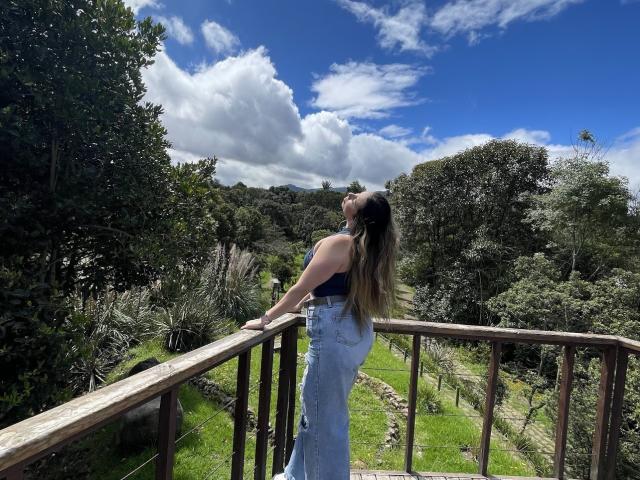 JennyLawry with long brown hair, wearing a blue tank top and high-waisted jeans, leans on a wooden balcony, looking up at a bright blue sky with fluffy clouds.