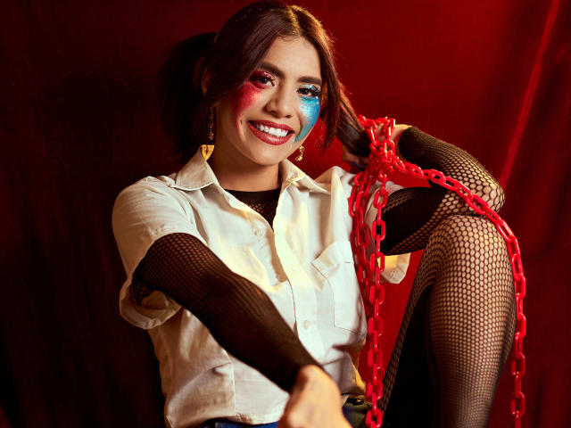 LucieMilner con la cara pintada de rojo y azul, camisa blanca, mangas de red negra y una cadena roja, sonriendo, con las piernas levantadas, mostrando la vulva cubierta con la red, cabello castaño oscuro, fondo rojo..