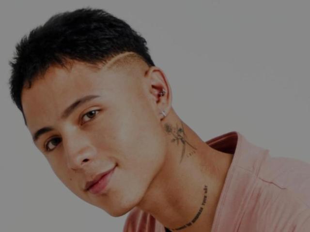 PiperrCry, un tipo de pelo corto con un corte degradado, con perforaciones en las orejas, un tatuaje pequeño en el cuello, una camisa rosa, ligeramente inclinado, sonriendo, piel clara, ojos marrones, sin barba, mirando hacia un lado..