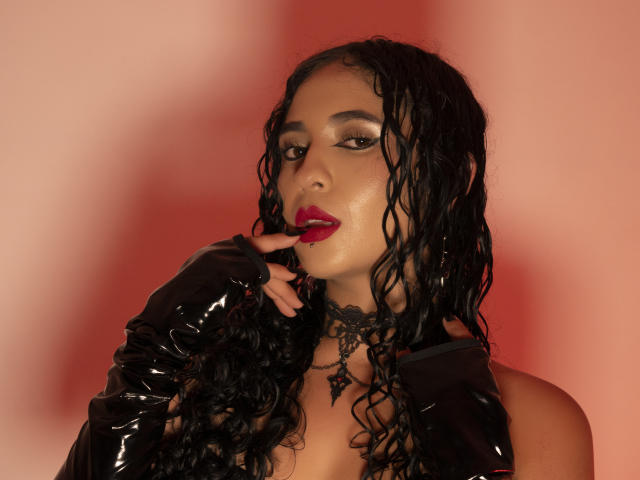 LilithStygian mit nassen, schwarzen Locken, glänzenden schwarzen Latexhandschuhen, rotem Lippenstift und einer schwarzen Spitzenkette, die mit ihrem Finger ihre Lippen berührt. Roter Hintergrund..
