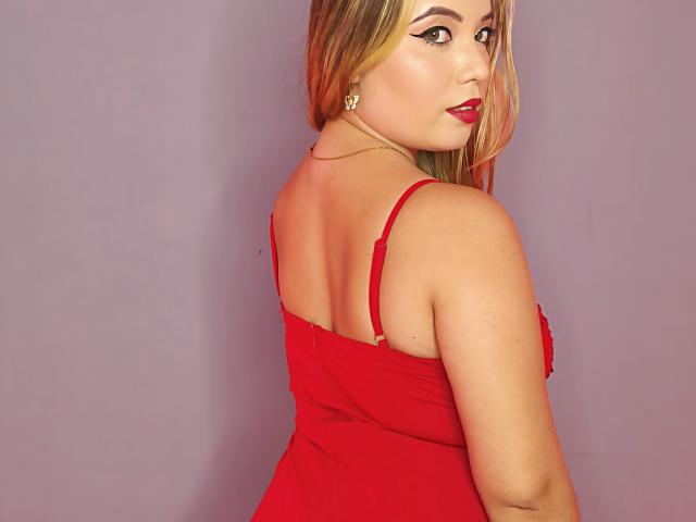 ZoeTobon69 has blonde hair, red lipstick, and is wearing a red dress with straps, showing her back and looking over her shoulder. Her tits are visible.