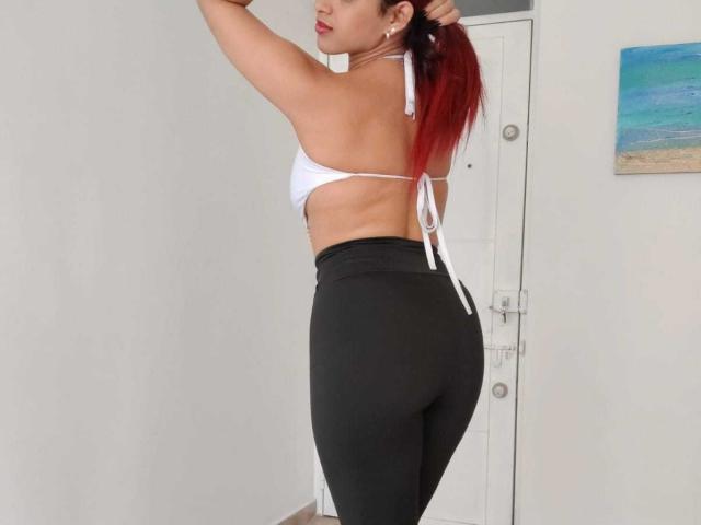 SweetieAlice69 with red ponytail, white halter bikini top, black high-waist leggings, showing her ass, standing side-profile, in a bright room.