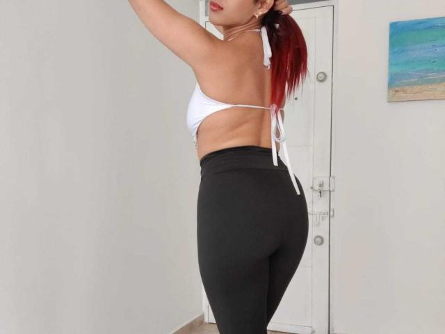 SweetieAlice69 with red ponytail, white bikini top, and black leggings, showing her ass, standing side profile, arm raised, white door in background, painting on right wall.