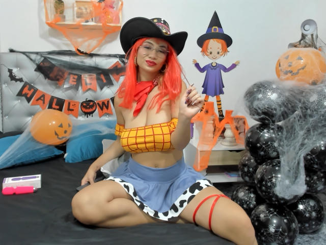MarianneBowe with bright red hair, wearing a black witch hat, yellow Spider-Man crop top, blue cow-print skirt, and red garter, sitting on a bed in Halloween cosplay.