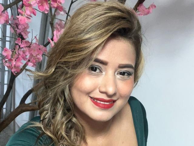 SecretGroupX has blonde, wavy hair, red lipstick, and is smiling at the camera with green eyes. She's wearing a green top and pink cherry blossoms are in the background.
