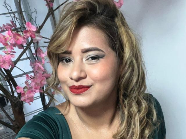 SecretGroupX has wavy blonde hair, red lipstick, and green eyes with smoky eyeliner. She's smiling with her head slightly tilted. She's wearing a green top.