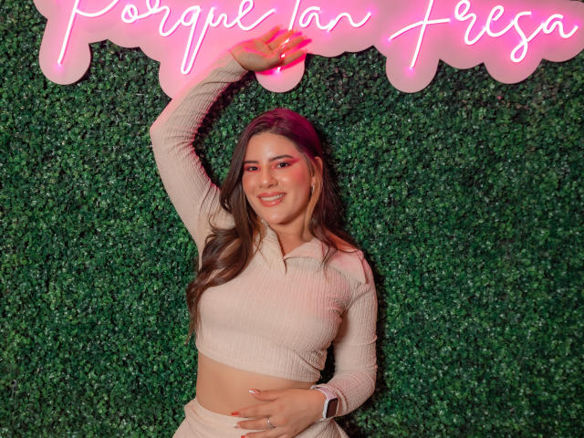 EmmaMack with long brown hair, wearing a pink ribbed crop top and beige pants, arm raised, smiling, green textured wall, neon pink sign above. Boobs, ass, tits visible.