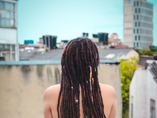 BeckyHuston, bare back visible, stands with long dark brown dreadlocks, some with small colorful beads, facing away. Urban rooftop in background, with blurred buildings and a clear blue sky. Textures of BeckyHuston's hair contrast with the smooth, slightly weathered rooftop. Colors: BeckyHuston's hair is dark, rooftop is beige, sky is blue. Composition: BeckyHuston centered, rooftop and sky blurred. Style: realistic photograph.