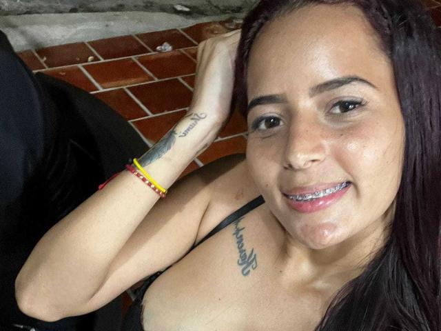 AdrianaMills with dark hair, smiling, showing a tattoo on her left forearm, another on her right chest, and a beaded bracelet, with visible white cum on her chin.