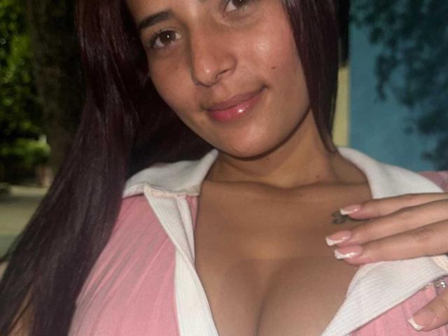 AdrianaMills with medium brown skin, dark brown hair, pink jacket, white collar, and hand on chest, showing her large, shiny, naked tits, smiling, with glossy pink lips.