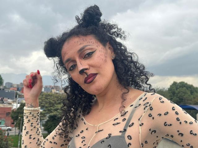 SeleneCollins with curly black hair in double buns, dark lipstick, leopard print top, face paint, and red nails, holding her hair up. Cloudy sky in background.