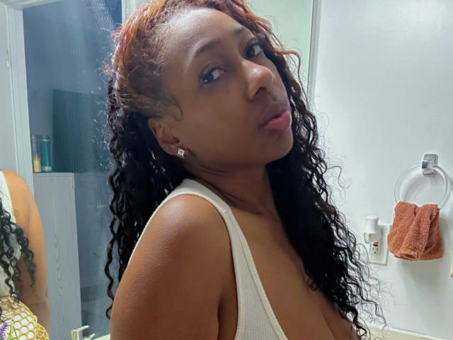 MadamGlam with dark curly hair, brown skin, and small tits in a white tank top, looking slightly to the side with pouty lips. Bathroom setting, towel ring in background.