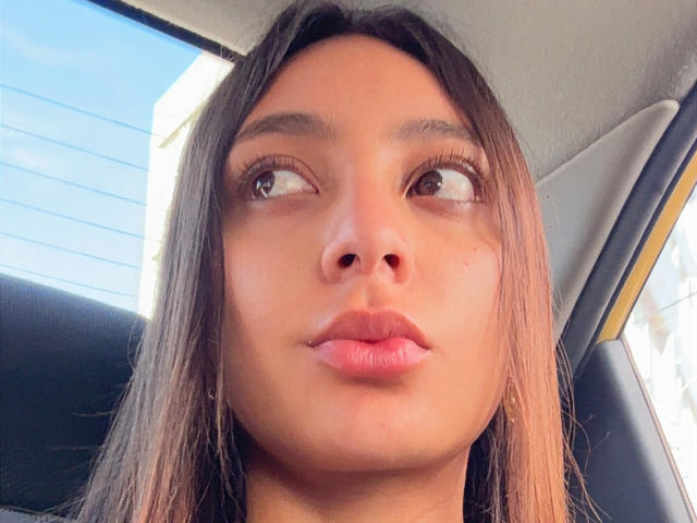 DianaFernandezz's face, brown eyes looking to the side, glossy pink lips, straight brown hair, tan skin, in a car, headrest visible, window to the right.