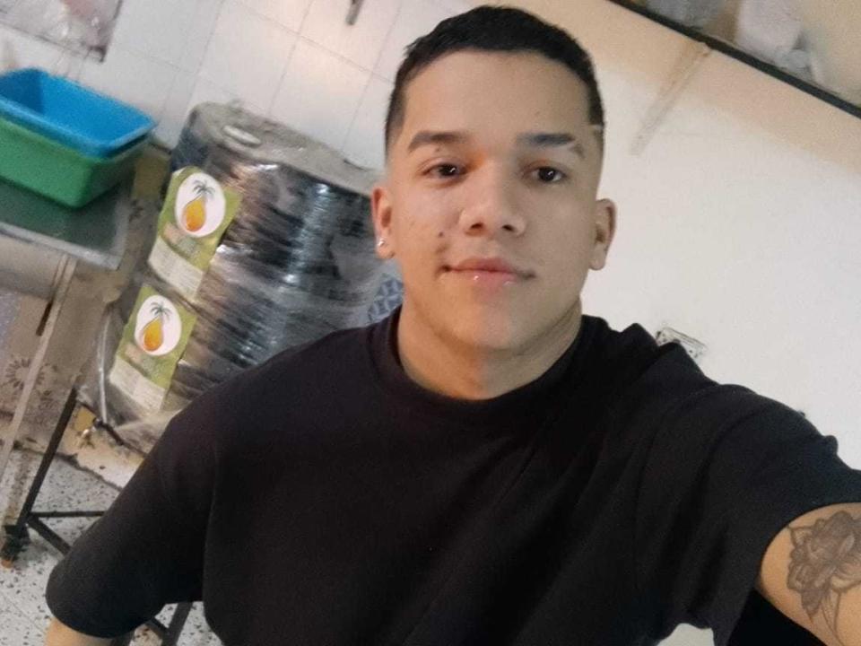 AndersMillan, short black hair, light skin, black t-shirt, small nose ring, tattoo on right arm, slight smile, standing in kitchen, stacks of plastic bags in background.