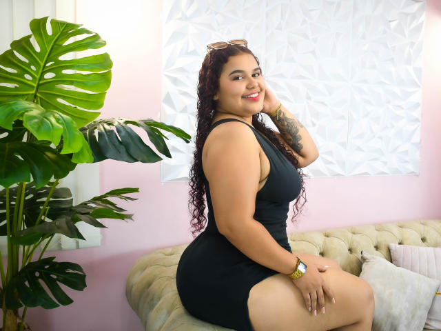 ClaireDolly with curly dark hair, wearing a black dress, sitting on a beige tufted sofa, smiling, showing her ass, with a green plant on the left and sunglasses on.