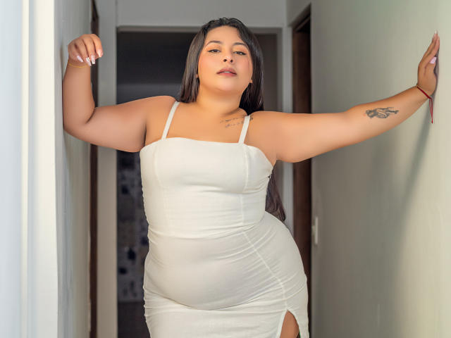 AllyCarson stands in a hallway, arms raised, wearing a white, form-fitting dress that shows her big ass and small tits. She has long black hair and a tattoo.