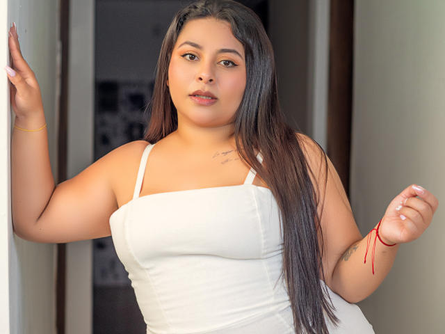 AllyCarson with long brown hair, white tank top, and red bracelet, standing, hand on wall, small tattoo on chest, brown eyes, slightly parted lips, curvy, medium tits.