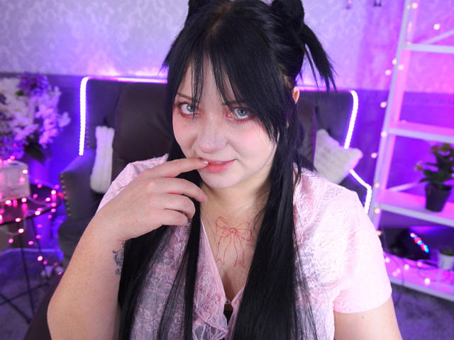 SweetLilu has pale skin and black hair in pigtails. She's wearing a pink lace top with a red ribbon design on the chest. Her blue eyes are slightly teary, and she's biting her finger. Her right hand has a small tattoo. The background is purple-lit with a black couch, white pillows, and a white shelf. The image has a casual, slightly provocative feel with soft, colorful lighting. The focus is on SweetLilu's facial expression and hair.