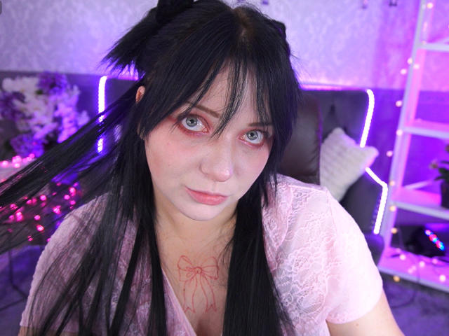 SweetLilu, con lunghi capelli neri, una maglietta di pizzo rosa e un tatuaggio rosso sul petto, guarda nella fotocamera con occhi azzurri, illuminazione viola e rosa, e un'espressione seria..
