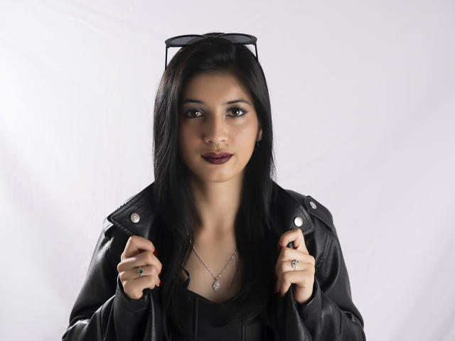 AlisKooper with long black hair, dark lipstick, wearing a black leather jacket, holding it with both hands, sunglasses on top, silver necklace, and multiple rings.